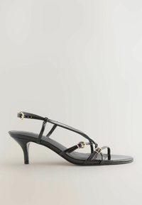 Sorte laklæder sandal med en slank hæl, udstyret med tynde stropper og guld hardware detaljer, herunder cirkulære ringelementer.