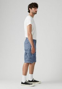 Man wearing white t-shirt, blue cargo shorts, white socks, and black sneakers standing sideways against a plain gray background.