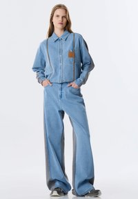 Lichtblauwe denim overhemd met een bruine leren patchzak, gecombineerd met lichtblauwe en grijze wijde broek. Moderne, ontspannen pasvorm.