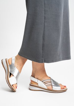 Silver metallic sandals with a cross strap design, featuring a light tan footbed and textured white sole. Worn with a gray midi dress.