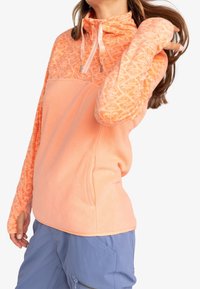 Femme portant un pull polaire à motifs pêche avec des trous pour les pouces, un col zippé et un pantalon cargo bleu, sur un fond blanc uni.