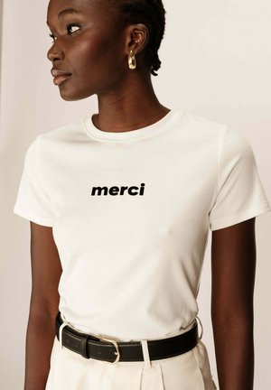 T-shirt en coton blanc avec le texte noir "merci", associé à une ceinture noire. Le t-shirt a un col rond et des manches courtes.