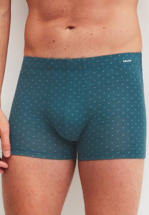Mannelijke torso gekleed in turquoise boxershorts met een klein stippenpatroon en het "CALIDA"-logo op de tailleband, staand tegen een effen achtergrond.