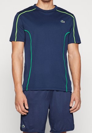 Mand iført navy blå sportstrøje og shortsæt med grøn piping og små krokodille-logoer på brystet og benet.