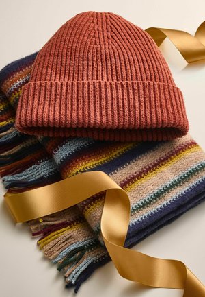 Un bonnet tricoté couleur rouille posé sur une écharpe rayée multicolore, avec des bords à franges et un ruban en satin doré. Texture textile douce.