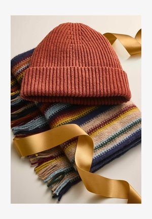 Rustrød strikket hue ovenpå et flerfarvet stribet tørklæde med frynser og et gyldent satinbånd. Blød tekstilstruktur.