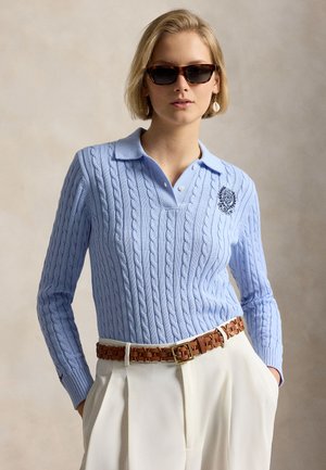 Kvinde iført solbriller med skildpaddemønster, lyseblå fiskebensstrikket polosweater med emblem, hvide bukser med høj talje og brunt flettet bælte, hænderne i lommerne.