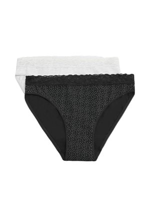 Mujer de pie con sujetador negro y ropa interior negra con detalles de encaje y pequeños puntos blancos, sobre un fondo liso y claro, mirando ligeramente de lado.