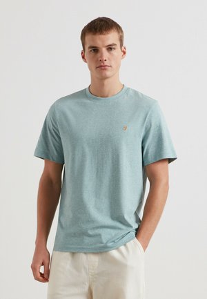 Jonge man draagt een lichtblauw T-shirt met korte mouwen en een klein geel logo op de borst, met crème kleurige broek, staande tegen een witte achtergrond.