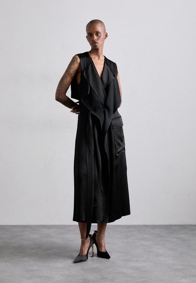 Black sleeveless dress with a draped front, featuring two side pockets and a textured finish. Paired with black pointed heels and ankle straps.