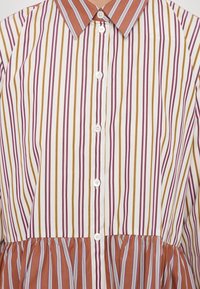 Chemise boutonnée à rayures verticales blanches, brunes, rouges et violettes, avec un col pointu et un détail de tissu froncé près de la taille.