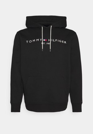 Schwarzer Hoodie mit weißen Kordeln, Fronttasche und dem Schriftzug „Tommy Hilfiger Est. 1985“ mit kleinem rot-weiß-blauem Flaggenlogo auf der Brust.