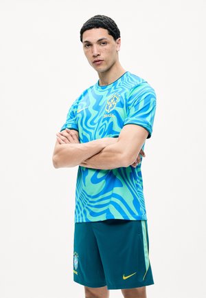 Młody mężczyzna ze skrzyżowanymi rękami, ubrany w niebiesko-zieloną koszulkę piłkarską Brazylii i ciemne szorty, stoi na jednolitym białym tle.