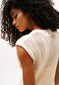 Top de punto sin mangas en color crema claro, con un diseño de malla, un cuello redondo y un pequeño detalle de marca en la parte trasera cerca del cuello.