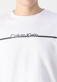 Weißes Baumwoll-T-Shirt mit einem schwarzen horizontalen Streifen und dem "Calvin Klein"-Logo in Kleinbuchstaben auf der Vorderseite; klassischer Rundhalsausschnitt.