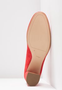 Escarpin en daim rouge avec semelle beige et petit talon bloc, posé semelle visible contre des murs blancs en angle.