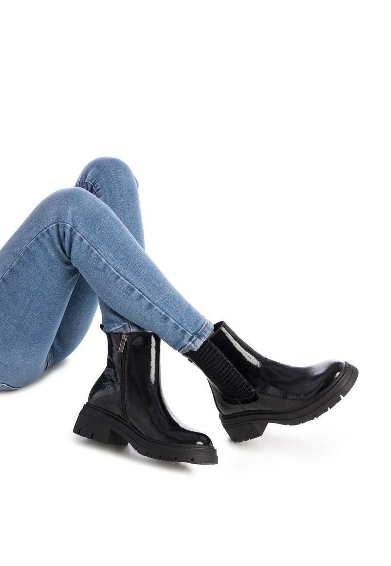 Botas de tobillo de charol negro con suela gruesa, cremallera lateral y paneles elásticos en los laterales, combinadas con vaqueros de denim azul.