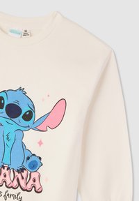 Felpa color crema con un personaggio dei cartoni animati blu con accenti rosa. Il design include testo e scintillii, con polsini a coste.