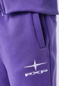 Plano detalle de unos pantalones de chándal morados con un bolsillo lateral y un logotipo bordado en blanco de PXP cerca del bolsillo.