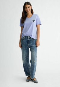 T-shirt lavande à col en V avec un imprimé floral, associé à un jean bleu déchiré avec des embellissements et une coupe décontractée. Ballet flats noires embellies.