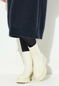 Stivali alla caviglia bianchi in pelle con suola spessa e pannelli laterali elasticizzati, abbinati a calze scure e a un vestito in maglia blu navy.