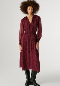 Burgundijas krāsas kleita ar riņķiem, garām caurspīdīgām piedurknēm, smockotu jostasvietu un midi garumu, apvienota ar melnām ceļgalu augstām zābakiem.