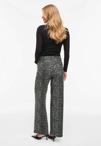 Pantalon à jambes larges en tissu à imprimé léopard vert foncé, avec une taille haute et des poches arrière, porté avec des chaussures à talons hauts noires.