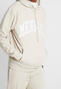 Beige huvtröja med "MENNACE" i vit text, känguruficka och justerbar dragsko. Mjuk textur med ribbade muddar.