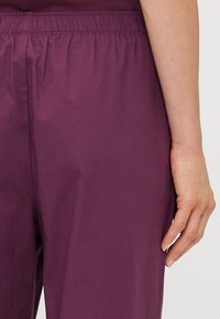 Pantalon violet décontracté avec une taille élastique, présentant une texture de tissu lisse et sans motifs ni accents visibles. Main positionnée sur le côté.
