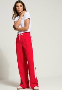 Rote weit geschnittene Hosen mit weißen Seitenstreifen und einem verstellbaren Bund, kombiniert mit einem schlichten weißen T-Shirt und hellen Schuhen.