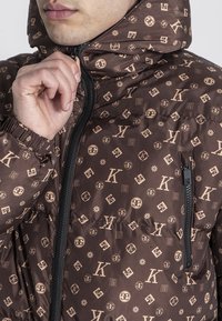 Hnedá puffa bunda so zvýraznenými logami v béžovej farbe, prešívaná textúra, vysoký golier a zipsová taška na hrudi.