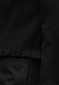 Pull en maille noire avec ourlet volanté et détail d'un pantalon en tissu noir assorti.