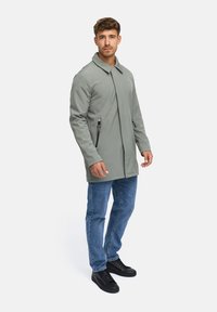 Grauer, weicher Mantel mit geradem Schnitt, großem Kragen und seitlichen Reißverschlüssen. Getragen mit blauen Jeans und schwarzen Sneakers. Einfarbiger Hintergrund.