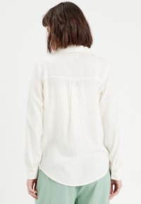 Blouse à manches longues, de couleur crème, avec un col à boutons, en tissu doux, et un ourlet légèrement arrondi. Le dos comporte des plis pour un détail supplémentaire.