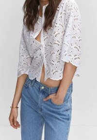 Blouse blanche en broderie anglaise avec manches courtes, ourlet festonné et fermeture à boutons sur le devant. Associée à un jean denim bleu clair.
