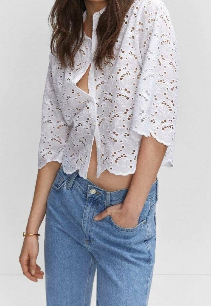 Blouse blanche en broderie anglaise avec manches courtes, ourlet festonné et fermeture à boutons sur le devant. Associée à un jean denim bleu clair.