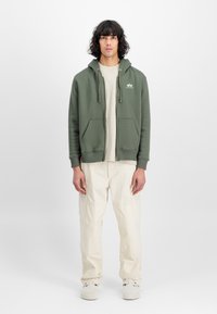 Ελιά χρώματος φούτερ με φερμουάρ, που διαθέτει μπροστινές τσέπες και ρυθμιζόμενα κορδόνια, φορεμένο πάνω από ένα μπεζ μπλούζα και σε συνδυασμό με ανοιχτό μπεζ παντελόνι.