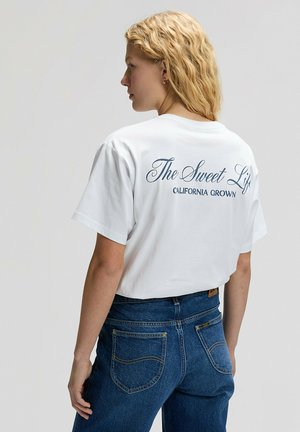 Młoda kobieta o kręconych blond włosach, ubrana w biały krótki t-shirt z napisem "The Sweet Life California Grown" oraz niebieskie dżinsy, stojąca bokiem.
