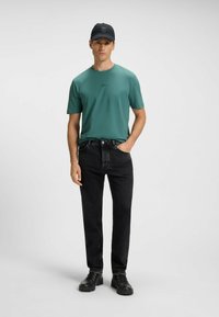 Dark black jeans with a straight fit, paired with a teal short-sleeve shirt. The model wears a black cap and sturdy black shoes.