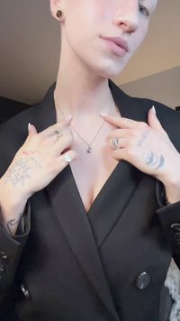 Colar com pendente de prata numa corrente fina, usado com um blazer preto. Tatuagens notáveis e vários anéis nas mãos, com unhas longas e de cor clara.