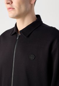 Schwarze Zip-Jacke mit Kragen, mit einem schwarzen geprägten Logo auf der linken Seite. Der Stoff hat eine glatte Textur.