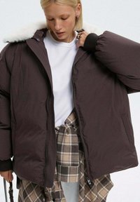 Brązowa kurtka puchowa z sztucznym futrem przy kołnierzu, zamek błyskawiczny oraz ściągacze przy rękawach, noszona na białej koszuli i w kratkowanych spodniach. Gładka tekstura.