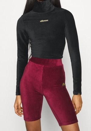 Femme portant un crop top noir à manches longues et col roulé, et un short de cyclisme en velours côtelé bordeaux taille haute.