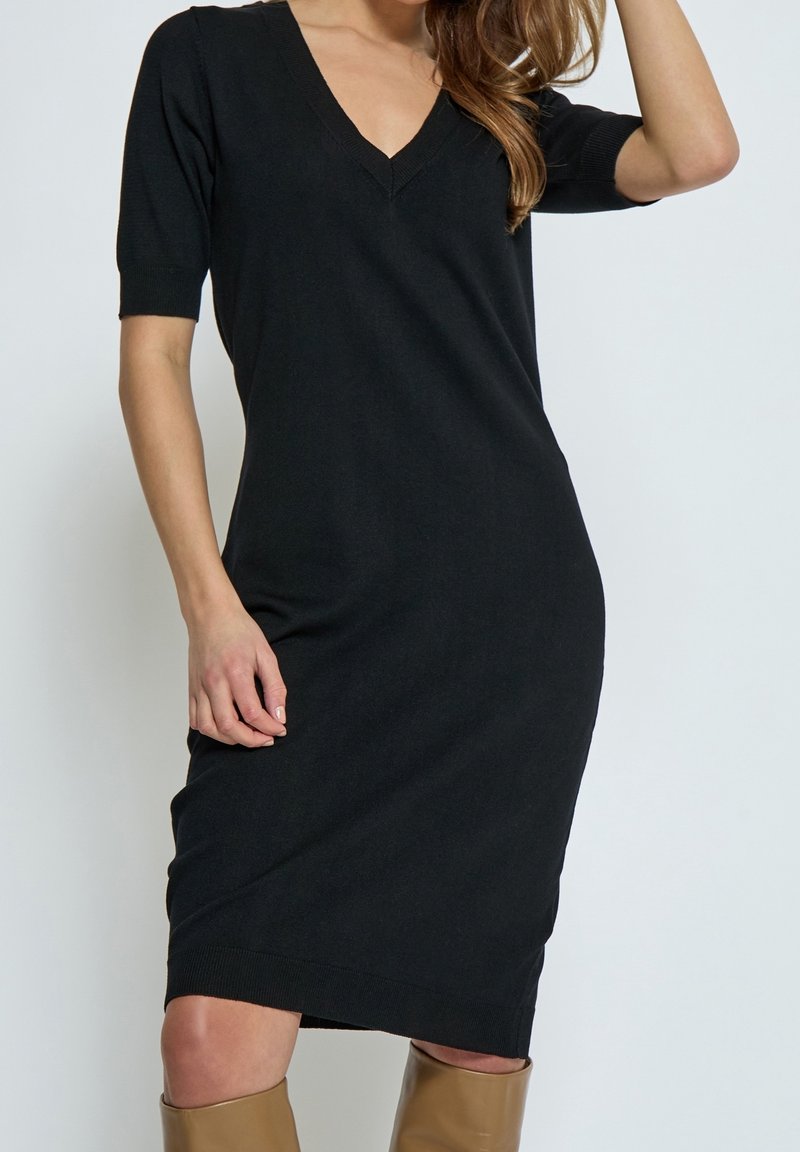 Femme portant une robe noire ajustée à col en V, longueur genou, manches courtes, avec des bottes hautes couleur beige sur fond uni.