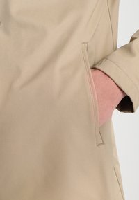 Giubbotto impermeabile beige con tasca laterale, texture liscia e vestibilità sartoriale. Le cuciture a vista e i dettagli di design sottili migliorano le funzionalità.