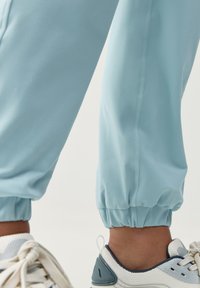 Světle modré joggingové kalhoty vyrobené z měkkého materiálu s elastickými manžetami a viditelným švem, kombinované s bílou teniskou s modrými akcenty.