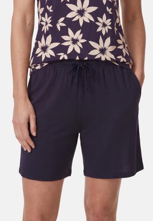 Frau trägt dunkelblaue Shorts mit Kordelzug und ein lila Oberteil mit Blumenmuster, Hände in den Taschen.