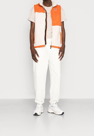 Wielobarwna kamizelka z pomarańczowego nylonu i kremowych paneli z polaru, z zamkiem z przodu i kieszenią na piersi, noszona na beżowej bluzce, z białymi spodniami i sportowymi butami.