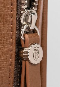 Tirante in pelle marrone con zip e un accento metallico, caratterizzato da un emblema circolare in rilievo e una finitura testurizzata sulla pelle.