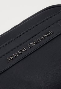 Borsa nera in ecopelle con superficie testurizzata e logo lucido "Armani Exchange" su una striscia rialzata. Design compatto ed elegante.
